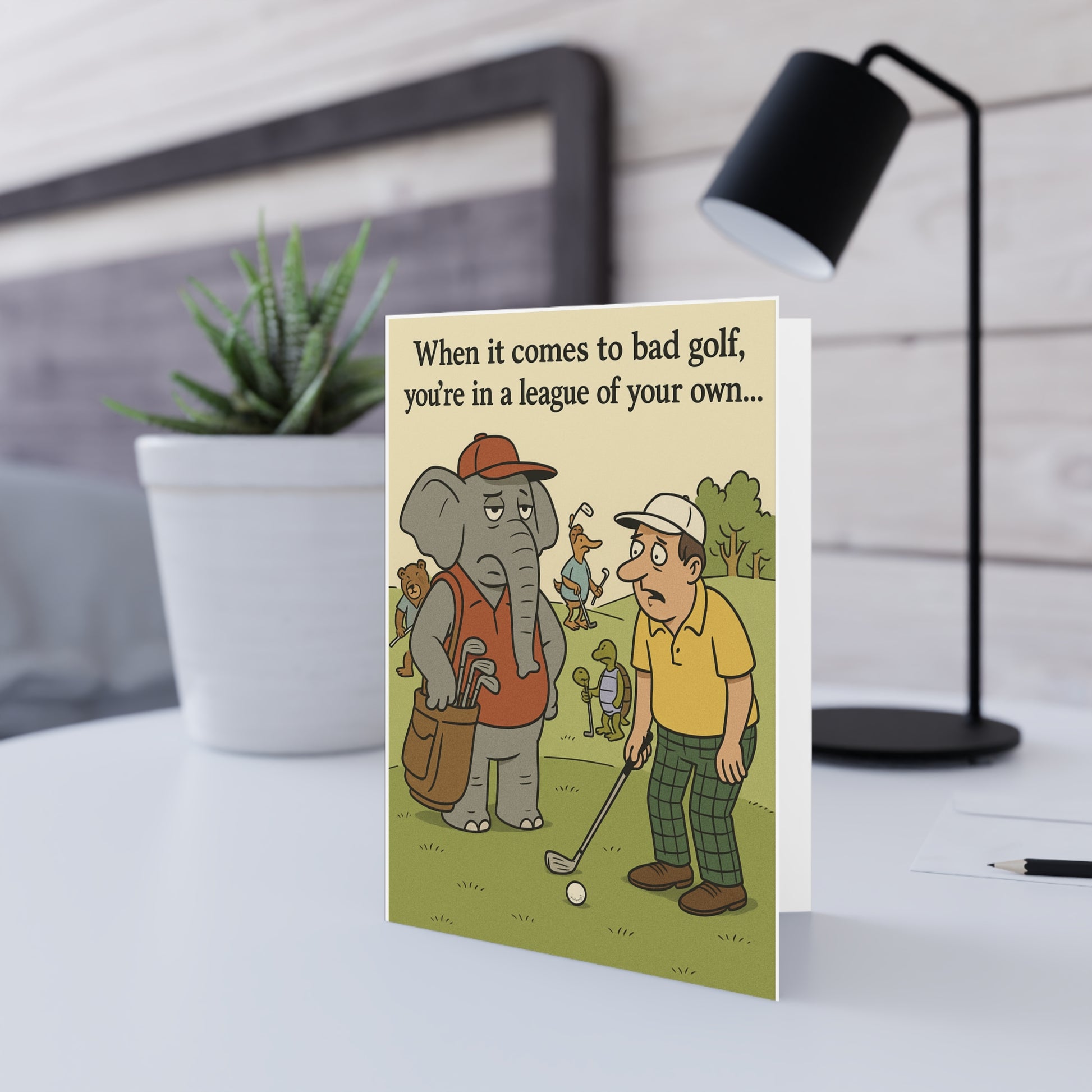 Greeting card with cartoon of a man and elephant playing golf on a desk with a lamp and plant in the background.